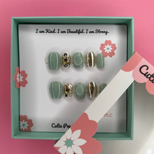 Green and Gold Sparkle press-on nails in a rich green base with sparkling gold accents and 3D bump embellishments, finished with a glossy shine – displayed on a Cutie Pop Nail Shop card.