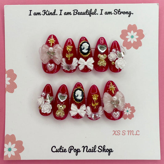 Victorian Shadows – Red press on nails by CutiePop Nail Shop featuring medium almond shaped nails in a deep red tone, adorned with delicate white rose art and ribbon details for a romantic gothic look.
