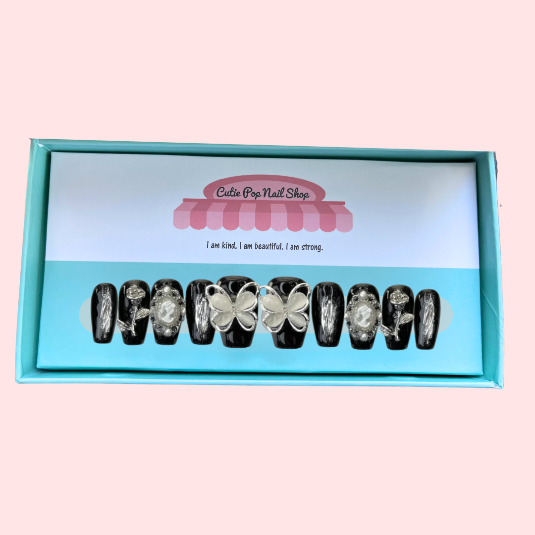 Black Butterfly press on nails by CutiePop Nail Shop featuring short length coffin shaped nails in a glossy jet black base. Several accent nails display clear backgrounds with bold black butterfly wing decals and metallic silver stud embellishments. Includes a full reusable kit: ten handmade nails, jelly adhesive tabs, mini nail file, and cuticle stick—offering a modern, edgy butterfly goth aesthetic.