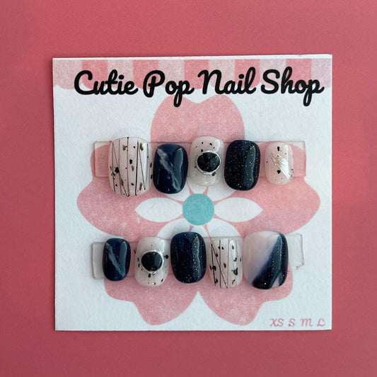 Abstract Gem press‑on nails by CutiePop Nail Shop featuring short-length squoval (square-oval) nails in a bold contrasting palette of glossy black and nude tones, accented by irregular abstract brush strokes and oversized metallic gemstone clusters. Includes reusable adhesive tabs, mini nail file, and a cuticle stick for full DIY application—delivering an edgy, artistic manicure with luxe shine.