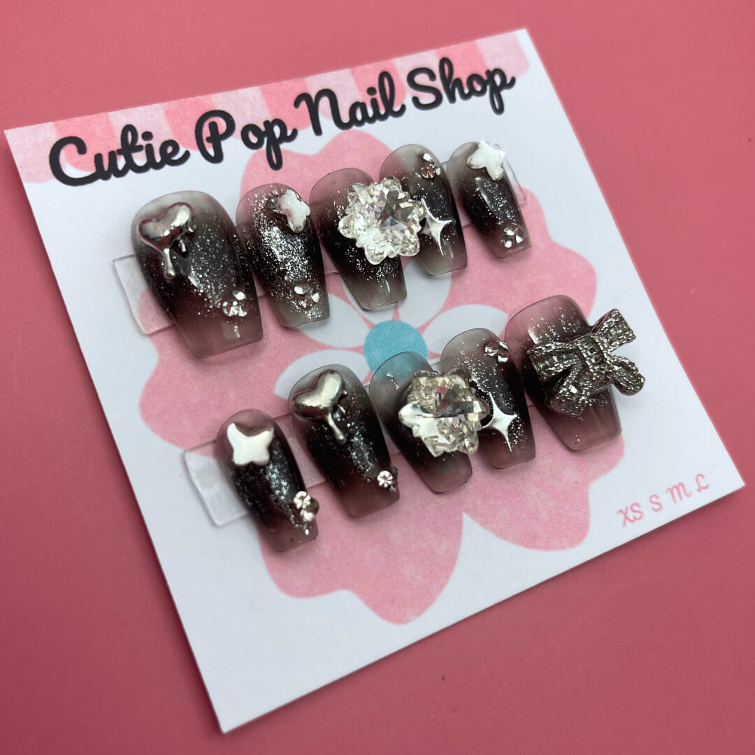 Black Gem Silver Glitter press‑on nails by CutiePop Nail Shop featuring medium-length coffin-shaped nails painted in a deep glossy black base, embellished with a sparkling silver glitter gradient and large gem clusters. The design includes multicolored rhinestones and metallic accents for a dramatic glam‑rock aesthetic. Arrives as a reusable kit—10 handmade press‑on nails, a sheet of jelly adhesive tabs, a mini nail file, and a cuticle stick.