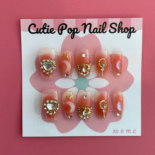 Blushing Moon press-on nails by CutiePop Nail Shop feature medium-length coffin-shaped nails in a blush-pink base, adorned with oversized starburst gem clusters, fine metallic glitter, and soft pearlescent shimmer. This dreamy set creates a romantic, glamorous statement look. Each kit includes 10 reusable handmade nails, jelly adhesive tabs, a mini nail file, and a cuticle stick for easy DIY application.