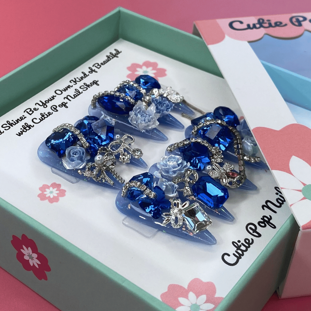 Bougie Bling 7 press on nails by CutiePop Nail Shop featuring long stiletto shaped nails with a vibrant royal blue glitter base adorned with oversized 3D gem clusters in matching blue and clear tones, complemented by sparkling silver rhinestones and metallic accents for a dazzling, high glam luxury statement manicure.