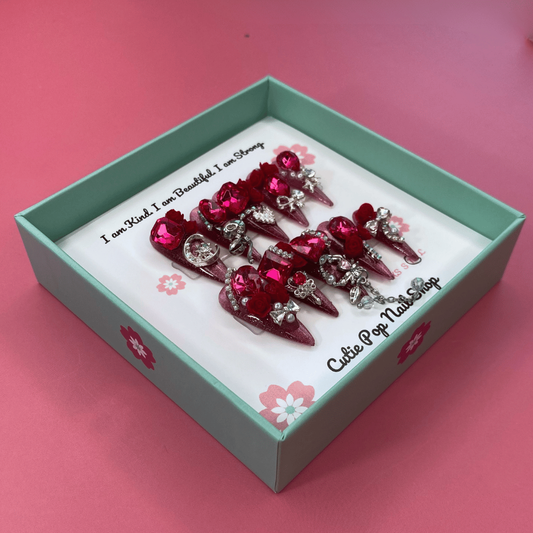 Bougie Bling 11 press on nails by CutiePop Nail Shop featuring long stiletto shaped nails with a bold wine red glitter base layered with luxurious oversized 3D gem clusters in deep jewel tones, accented with sparkling rhinestones and metallic detailing. Hand sculpted for a dramatic, upscale statement mani.