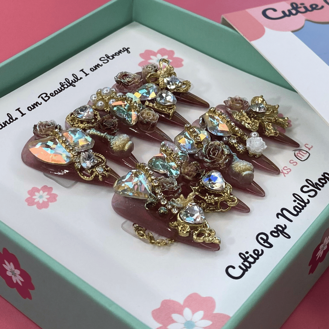 Bougie Bling 12 press on nails by CutiePop Nail Shop featuring long stiletto shaped nails with a glamorous deep dark burgundy or smoky wine toned glitter base. Lavish oversized 3D gem clusters in jewel tones such as ruby and crystal are complemented by sparkling rhinestones and metallic accents—arranged in an opulent, high impact luxury statement manicure.