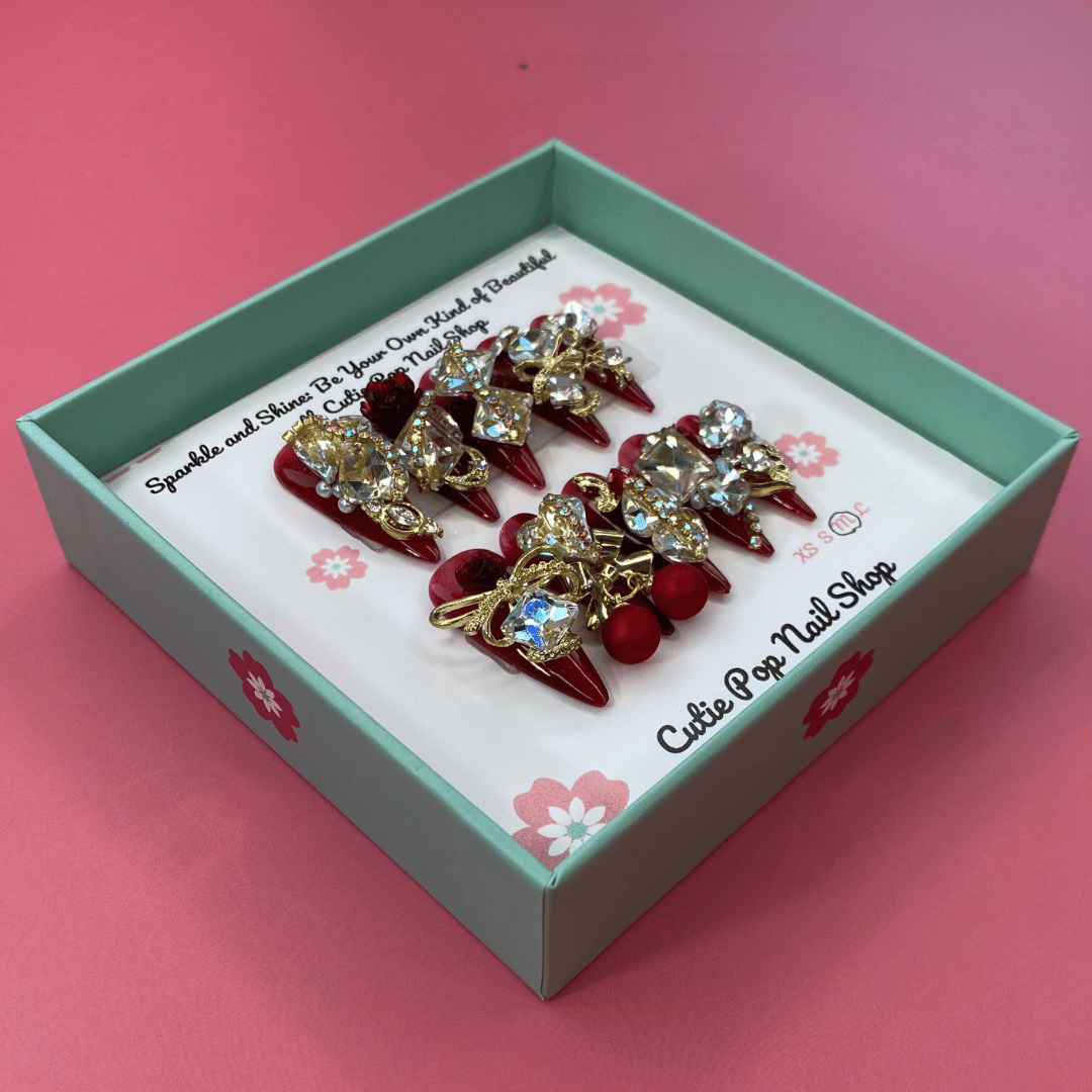Bougie Bling 13 press on nails by CutiePop Nail Shop featuring long stiletto shaped nails with a rich deep burgundy red or wine toned glitter base. Lavish oversized 3D gem clusters in jewel tones including ruby and crystal are accentuated with sparkling rhinestones and metallic highlights for a dramatic, high end glitzy manicure.