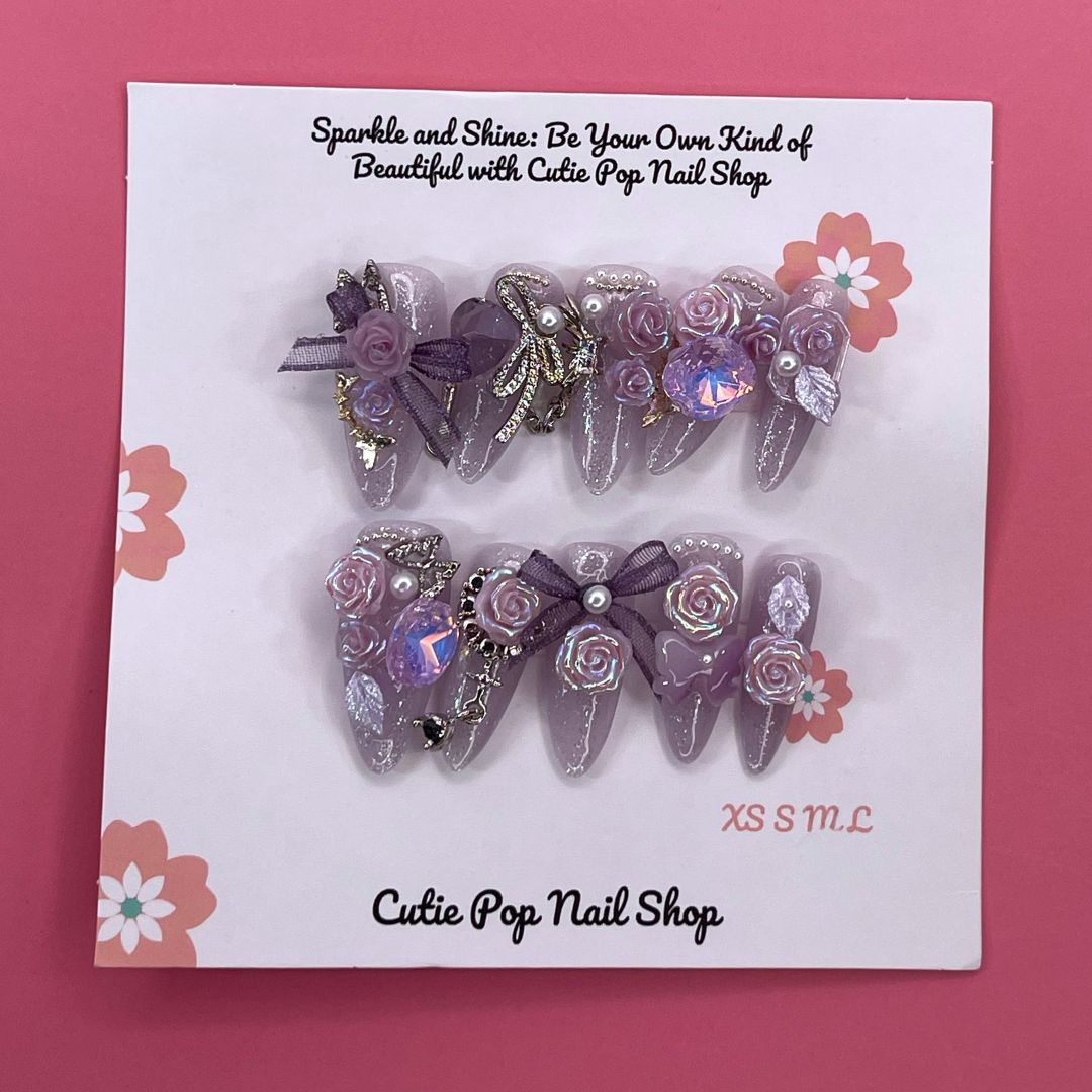 Bougie Bling 16 press-on nails by CutiePop Nail Shop feature medium-length almond-shaped nails in a shimmering lavender-purple glitter base, adorned with ornate 3D gem clusters and delicate rhinestone accents. Each nail is meticulously hand-sculpted to create a luxurious, playful-glam statement manicure. Includes 10 reusable nails, jelly adhesive tabs, a mini nail file, and a cuticle stick.