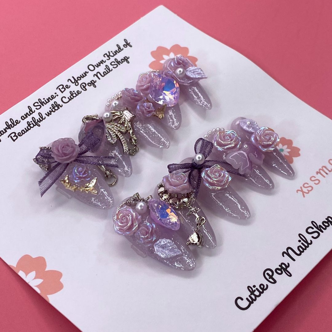 Bougie Bling 16 press-on nails by CutiePop Nail Shop feature medium-length almond-shaped nails in a shimmering lavender-purple glitter base, adorned with ornate 3D gem clusters and delicate rhinestone accents. Each nail is meticulously hand-sculpted to create a luxurious, playful-glam statement manicure. Includes 10 reusable nails, jelly adhesive tabs, a mini nail file, and a cuticle stick.