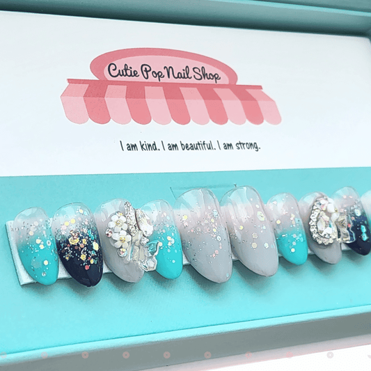 Fairytale press‑on nails by CutiePop Nail Shop featuring medium-length almond and round-shaped nails in a dreamy turquoise-to-navy gradient. These whimsical nails are accented with iridescent resin butterfly charms, holographic sequin glitter, and shimmering tones that evoke a magical wonderland vibe. Each set includes 10 reusable handmade nails, jelly adhesive tabs, a mini nail file, and a cuticle stick—perfect for an elegant yet playful fairy-tale inspired manicure.