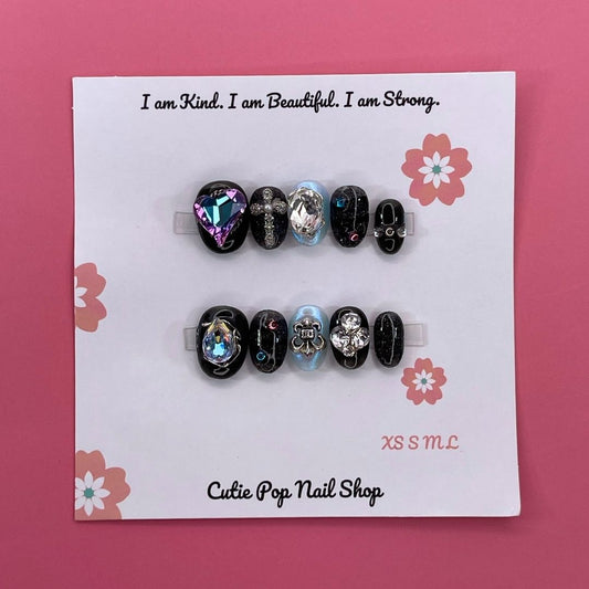 Cosmic Goth press on nails by CutiePop Nail Shop featuring medium round shaped nails in black and purple tones with celestial designs like stars and moons, accented with metallic or glittery finishes for a dark, dreamy aesthetic.