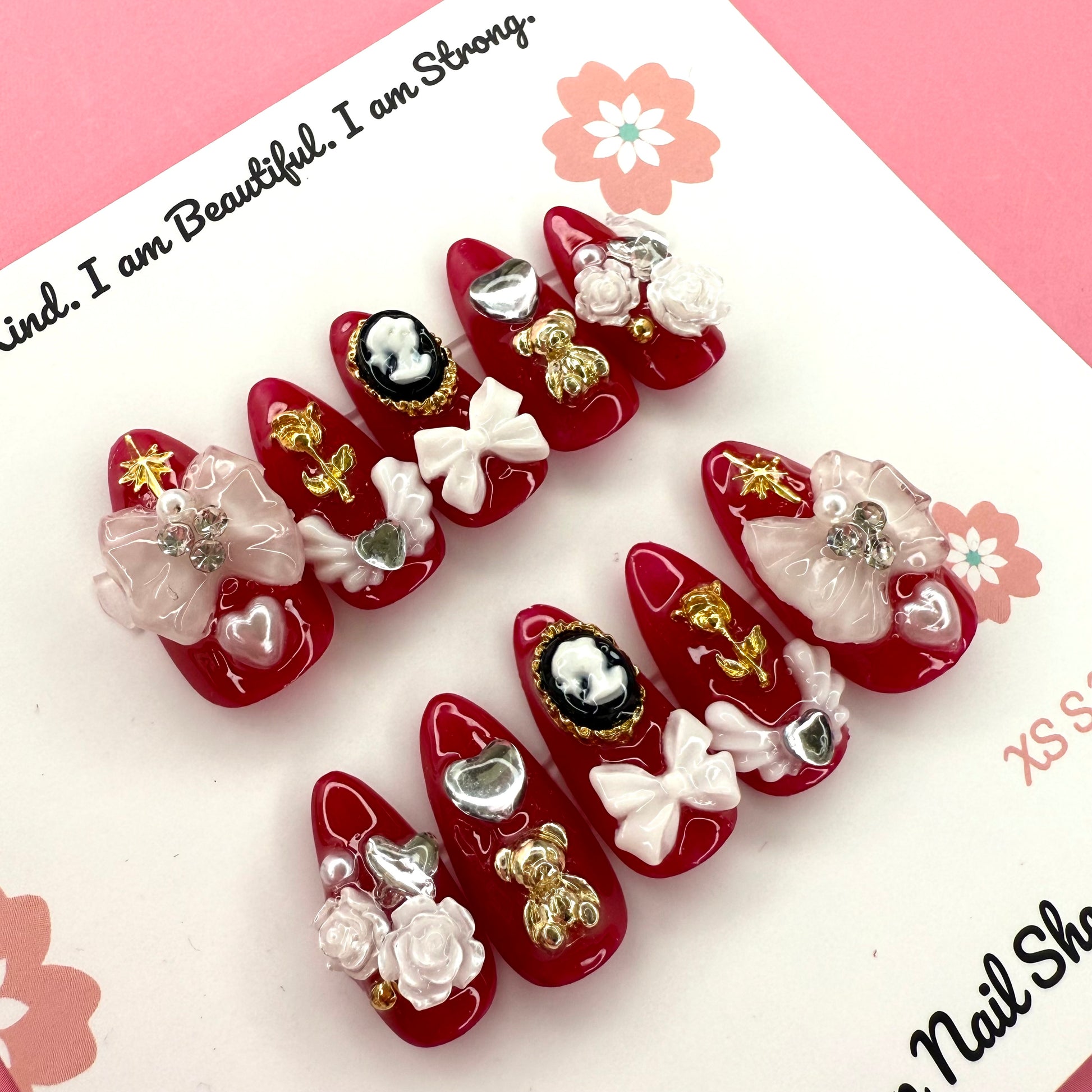 Victorian Shadows – Red press on nails by CutiePop Nail Shop featuring medium almond shaped nails in a deep red tone, adorned with delicate white rose art and ribbon details for a romantic gothic look.