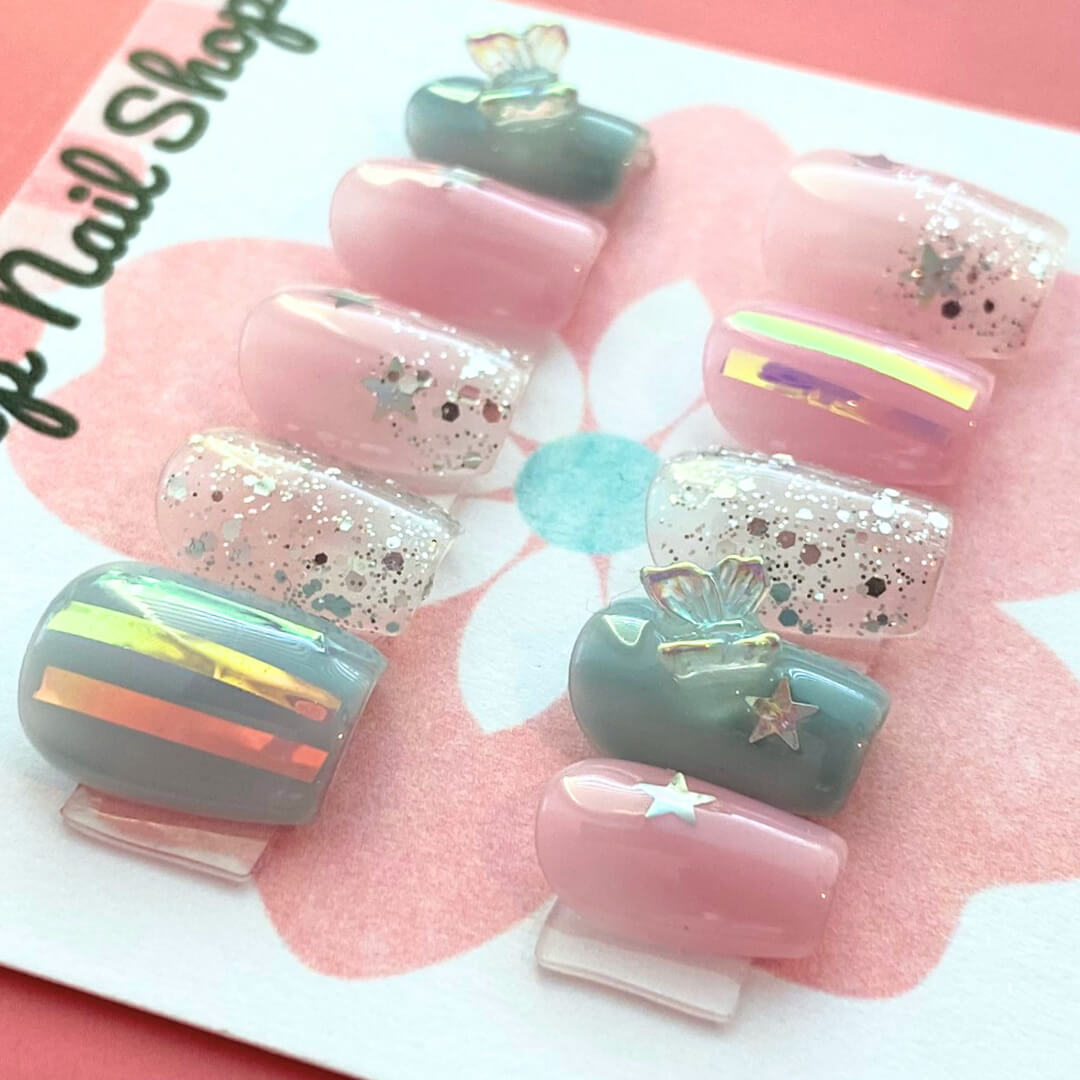 Dreamy Butterfly press‑on nails by CutiePop Nail Shop featuring coffin-shaped extra short nails in soft pastel pinks and blues, accented with hand-painted butterfly wing motifs, metallic foil shimmer, and delicate glitter sparkle. The set includes reusable press-on tabs, a mini nail file, and a cuticle stick—offering a fantasy-inspired, playful yet elegant manicure.