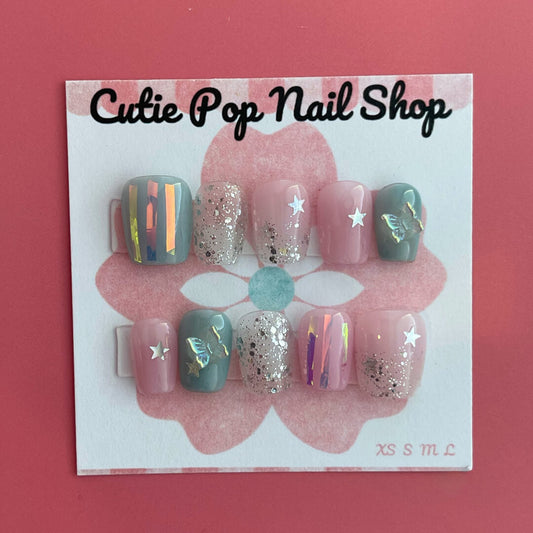 Dreamy Butterfly press‑on nails by CutiePop Nail Shop featuring coffin-shaped extra short nails in soft pastel pinks and blues, accented with hand-painted butterfly wing motifs, metallic foil shimmer, and delicate glitter sparkle. The set includes reusable press-on tabs, a mini nail file, and a cuticle stick—offering a fantasy-inspired, playful yet elegant manicure.