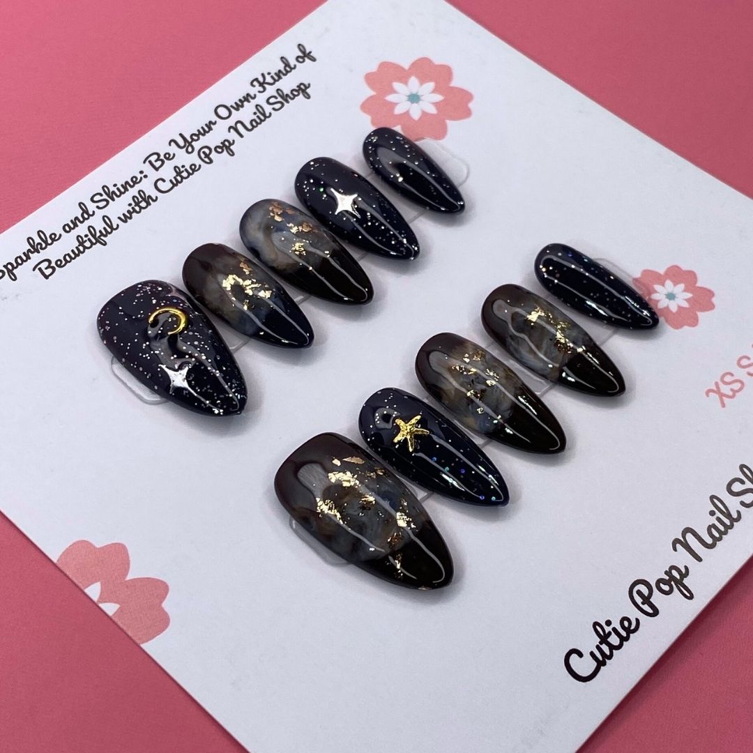 Galaxy Stars press on nails by CutiePop Nail Shop featuring medium almond shaped nails in deep navy and black tones, adorned with shimmering star, moon, and cosmic accents for a bold, celestial-inspired look.