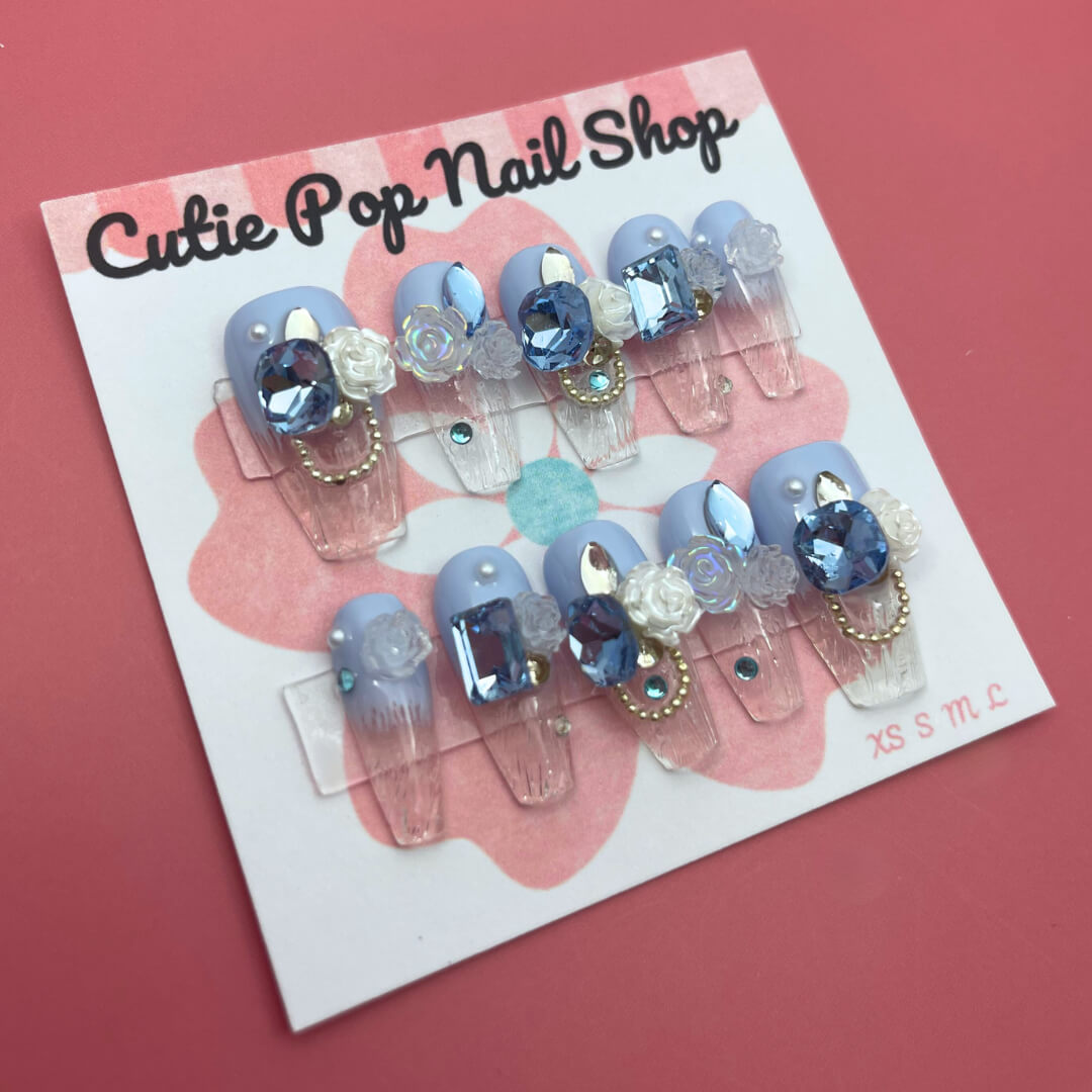 Elegant Rose (Blue) press on nails by CutiePop Nail Shop featuring medium coffin shaped nails in a rich sapphire blue or teal base, decorated with finely hand painted rose motifs in soft blush or pale pink tones. Accented with subtle metallic shimmer and tiny pearl or crystal details at the rose centers, finished with a glossy gel top coat. The set includes 10 reusable handmade nails, jelly adhesive tabs, a mini nail file, and a cuticle stick—designed for a refined, floral luxe manicure.
