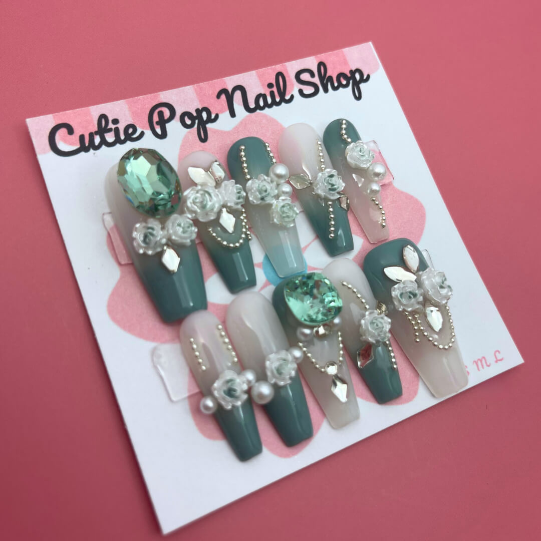 Elegant Rose (Green) press‑on nails by CutiePop Nail Shop featuring long coffin-shaped nails in a rich emerald-green base accented with intricately hand-painted delicate rose motifs in soft beige or cream hues. Embellished with subtle metallic shimmer and tiny pearl or rhinestone details at the center of each rose, finished with glossy gel polish for a refined, elegant floral manicure. Includes 10 handmade reusable nails, jelly adhesive tabs, a mini nail file, and cuticle stick for a full DIY kit.