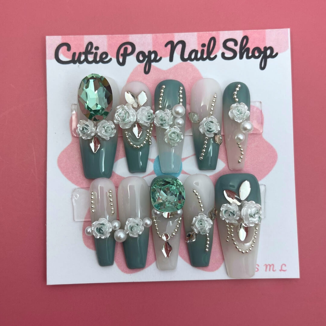 Elegant Rose (Green) press‑on nails by CutiePop Nail Shop featuring long coffin-shaped nails in a rich emerald-green base accented with intricately hand-painted delicate rose motifs in soft beige or cream hues. Embellished with subtle metallic shimmer and tiny pearl or rhinestone details at the center of each rose, finished with glossy gel polish for a refined, elegant floral manicure. Includes 10 handmade reusable nails, jelly adhesive tabs, a mini nail file, and cuticle stick for a full DIY kit.
