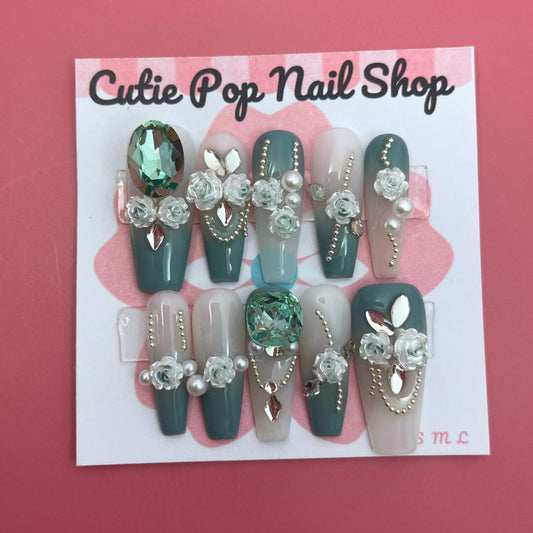 Elegant Rose (Green) press‑on nails by CutiePop Nail Shop featuring long coffin-shaped nails in a rich emerald-green base accented with intricately hand-painted delicate rose motifs in soft beige or cream hues. Embellished with subtle metallic shimmer and tiny pearl or rhinestone details at the center of each rose, finished with glossy gel polish for a refined, elegant floral manicure. Includes 10 handmade reusable nails, jelly adhesive tabs, a mini nail file, and cuticle stick for a full DIY kit.
