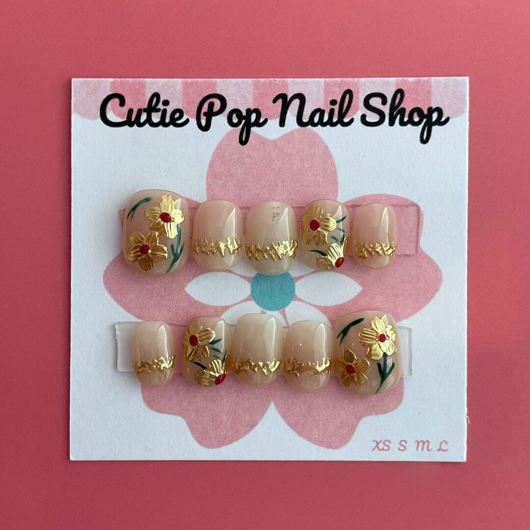 Golden Flower press‑on nails by CutiePop Nail Shop featuring extra-short squoval nails with a translucent base layered in reflective gold chrome flower embellishments. Includes a full reusable kit: adhesive tabs, mini nail file, and cuticle stick—offering a delicate, glimmering floral statement with a subtle luxurious sheen.
