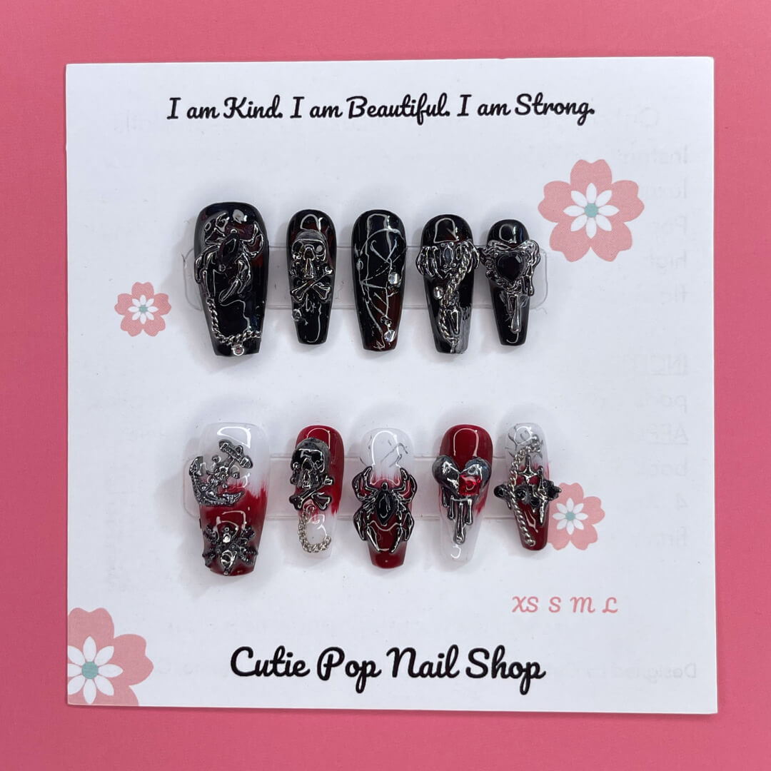 Gothic Delight press on nails by CutiePop Nail Shop featuring long coffin shaped nails painted in glossy jet black polish, accented with dark red duochrome shimmer and intricate gothic motifs like crosses or rose patterns, finished with subtle metallic or glitter highlights for a dramatic, dark romantic aesthetic.