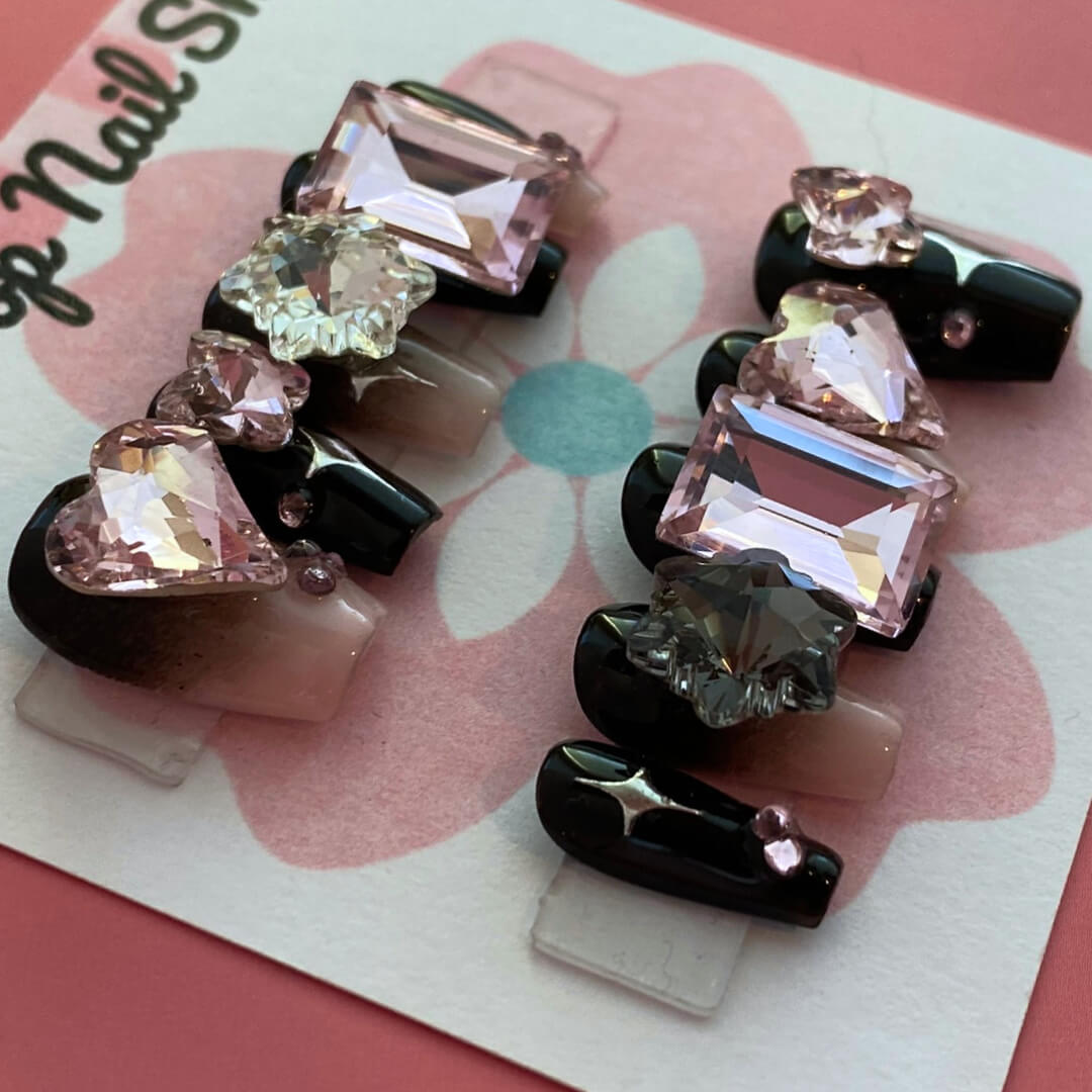 Gothic Girly press‑on nails by CutiePop Nail Shop featuring medium-length coffin-shaped nails painted in glossy black with alternating muted pink accent nails. Adorned with edgy gothic motifs—such as skulls, crosses, and mini bows—plus glittery accents and metallic studs, the design balances feminine charm with dark, rebellious elegance. The set arrives with reusable adhesive tabs, a mini nail file, and a cuticle stick for a complete application kit.