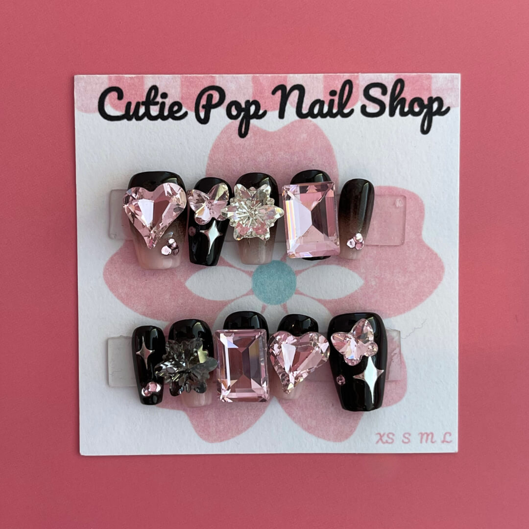 Gothic Girly press‑on nails by CutiePop Nail Shop featuring medium-length coffin-shaped nails painted in glossy black with alternating muted pink accent nails. Adorned with edgy gothic motifs—such as skulls, crosses, and mini bows—plus glittery accents and metallic studs, the design balances feminine charm with dark, rebellious elegance. The set arrives with reusable adhesive tabs, a mini nail file, and a cuticle stick for a complete application kit.
