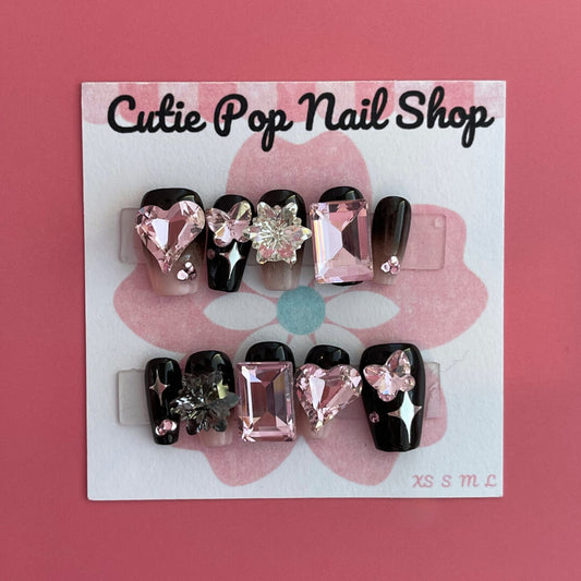Gothic Girly press‑on nails by CutiePop Nail Shop featuring medium-length coffin-shaped nails painted in glossy black with alternating muted pink accent nails. Adorned with edgy gothic motifs—such as skulls, crosses, and mini bows—plus glittery accents and metallic studs, the design balances feminine charm with dark, rebellious elegance. The set arrives with reusable adhesive tabs, a mini nail file, and a cuticle stick for a complete application kit.