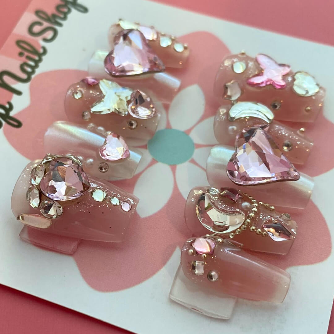 Magical Rhinestones press‑on nails by CutiePop Nail Shop featuring short to medium-length coffin-shaped nails in a soft blush-nude base, each adorned with oversized, iridescent rhinestone clusters forming floral and starburst patterns. The design sparkles with multi-faceted gem accents and metallic highlights for a luxe, high-bling aesthetic. The kit includes 10 reusable handmade nails, jelly adhesive tabs, a mini nail file, and a cuticle stick—perfect for glamorous DIY application.