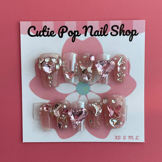 Magical Rhinestones press‑on nails by CutiePop Nail Shop featuring short to medium-length coffin-shaped nails in a soft blush-nude base, each adorned with oversized, iridescent rhinestone clusters forming floral and starburst patterns. The design sparkles with multi-faceted gem accents and metallic highlights for a luxe, high-bling aesthetic. The kit includes 10 reusable handmade nails, jelly adhesive tabs, a mini nail file, and a cuticle stick—perfect for glamorous DIY application.