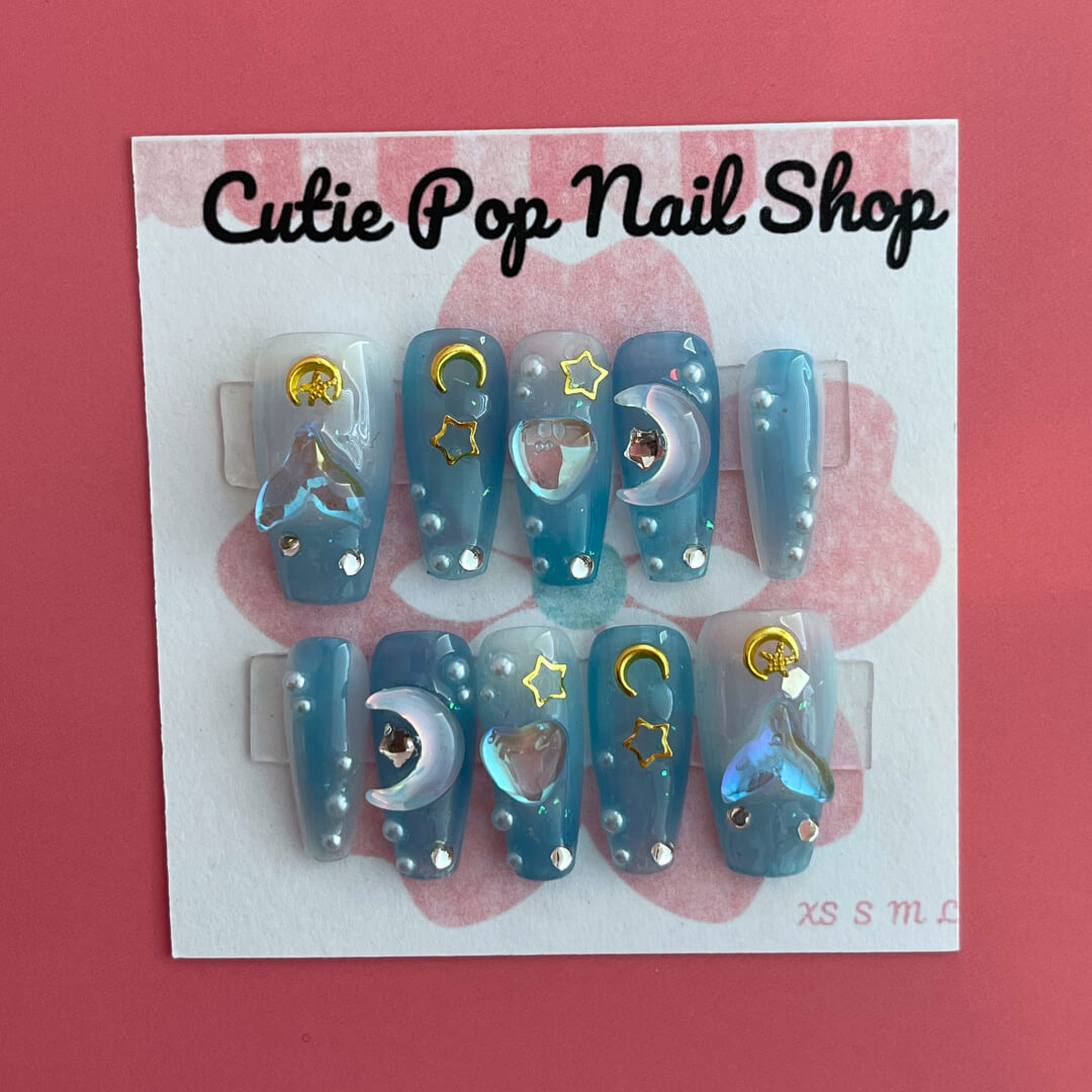 Mermaid Night press‑on nails by CutiePop Nail Shop featuring medium-length coffin-shaped nails in a gradient of deep teal and ocean blue hues, accented with iridescent shimmer, metallic starfish and shell charms, and subtle glitter specks for a mystical under-the-sea fantasy. Includes a full kit with reusable adhesive tabs, a mini nail file, and a cuticle stick—perfect for a dreamy mermaid-inspired manicure.