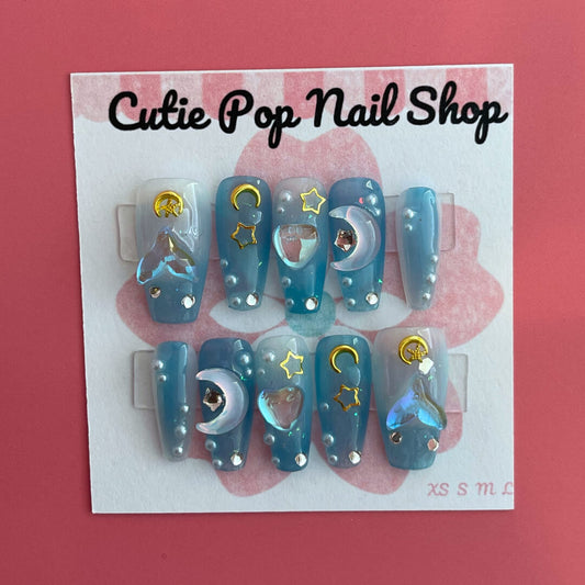 Mermaid Night press‑on nails by CutiePop Nail Shop featuring medium-length coffin-shaped nails in a gradient of deep teal and ocean blue hues, accented with iridescent shimmer, metallic starfish and shell charms, and subtle glitter specks for a mystical under-the-sea fantasy. Includes a full kit with reusable adhesive tabs, a mini nail file, and a cuticle stick—perfect for a dreamy mermaid-inspired manicure.
