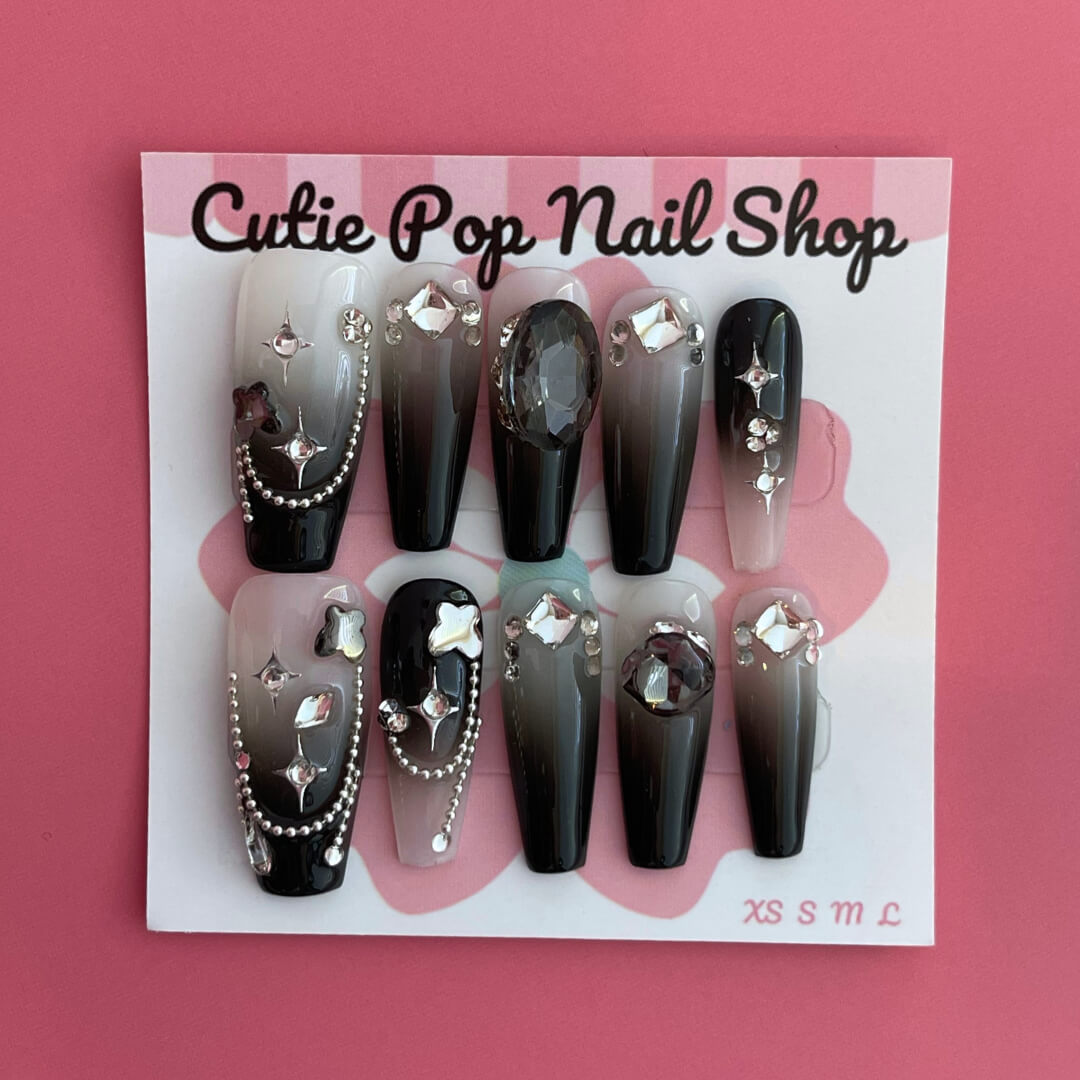 Midnight Whisper press-on nails by CutiePop Nail Shop featuring medium-length coffin-shaped nails with a deep black base, silvery metallic swirl motifs, and star-like shimmer accents. Designed in a limited-edition celestial gothic style, this set captures whispery galaxy vibes with an elegant, mysterious flair. Each kit includes 10 reusable handmade nails, jelly adhesive tabs, a mini nail file, and a cuticle stick for easy DIY application.
