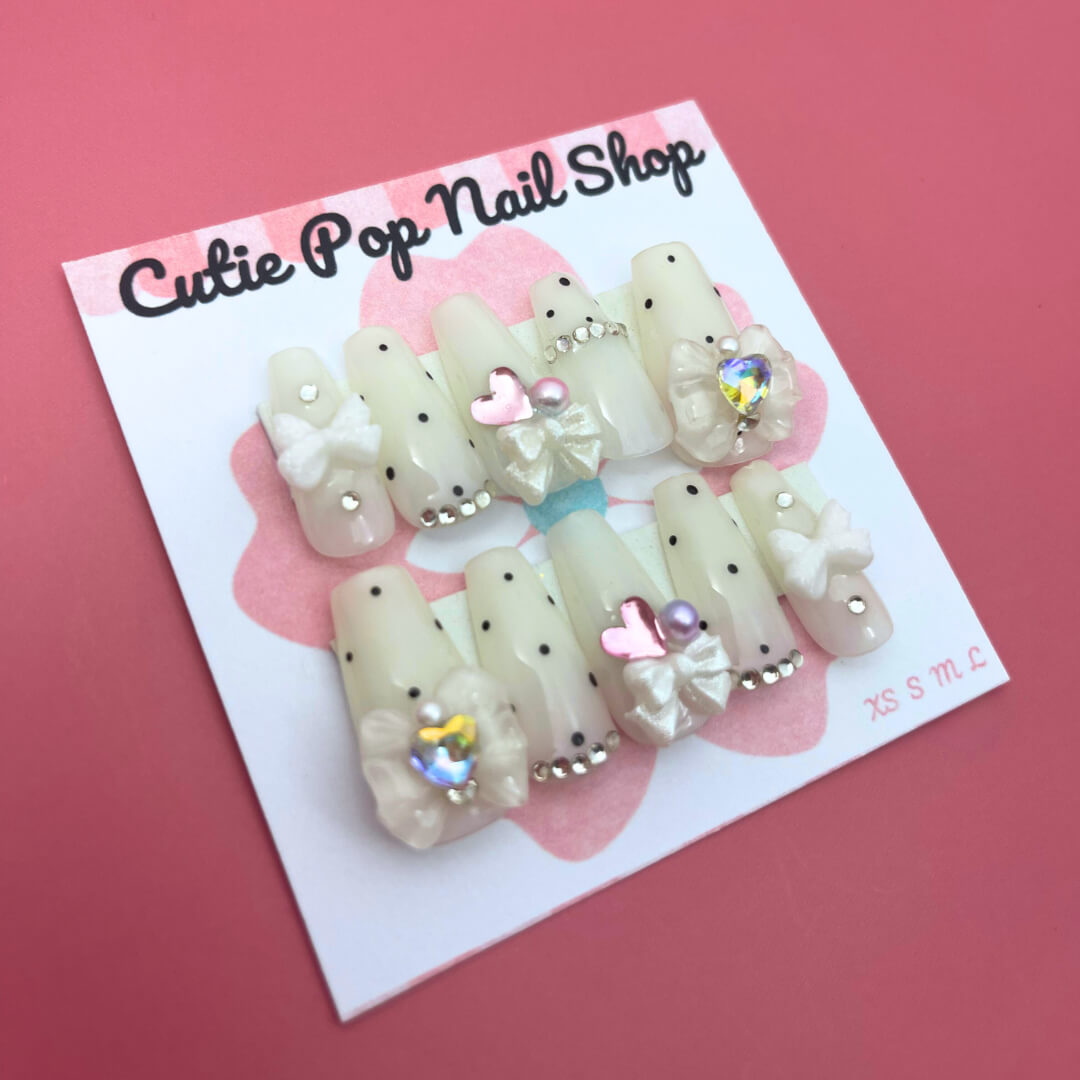 Polka Dot Pink press‑on nails by CutiePop Nail Shop featuring medium‑length coffin-shaped nails in a vibrant bubblegum-pink base with crisp white polka dots evenly spaced across each nail. The glossy finish enhances the playful, retro aesthetic. Includes a full reusable kit with ten handmade nails, jelly adhesive tabs, a mini nail file, and a cuticle stick—perfect for a fun, vintage-inspired manicure.