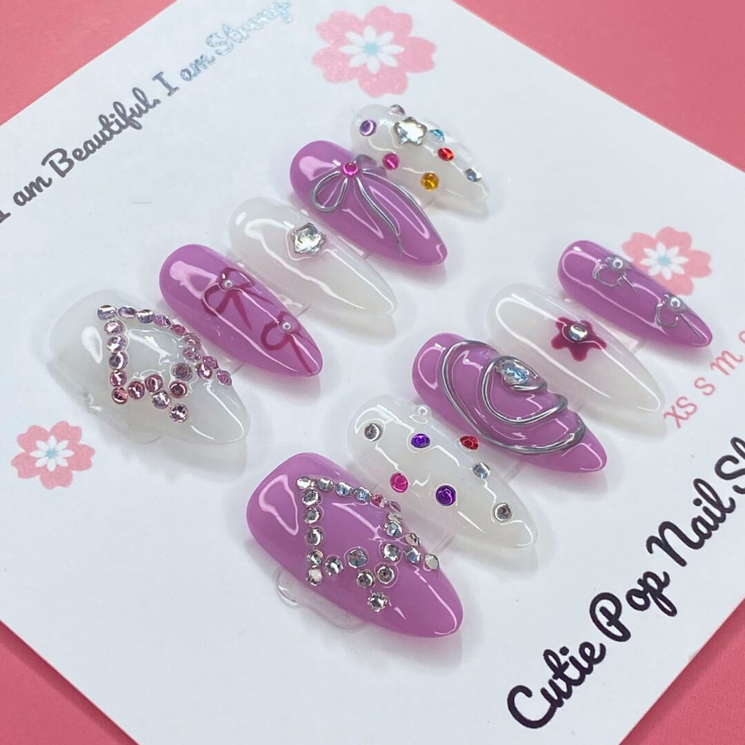 Purple Glamour press-on nails by CutiePop Nail Shop featuring medium-length almond-shaped nails in a mix of deep purple and crisp white tones. Accented with colorful gem clusters, embossed ribbon, star, and flower motifs, each nail is finished with a glossy shine for a chic and feminine glam look.