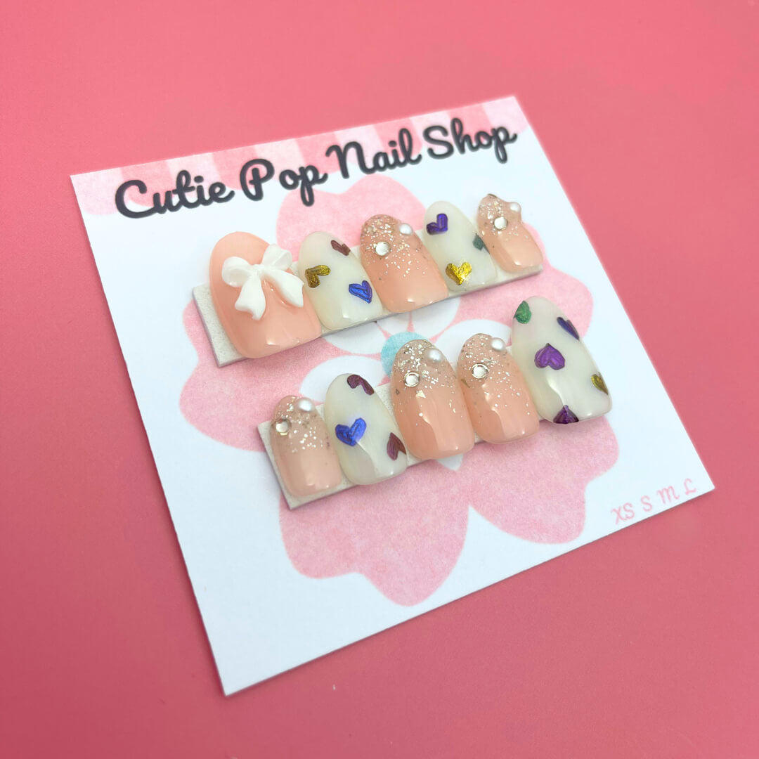 Rainbow Hearts press‑on nails by CutiePop Nail Shop featuring medium‑length round-shaped nails in a soft translucent pastel base, each adorned with hand-painted rainbow-colored heart motifs against a dreamy glossy finish. The nails come as a reusable kit—including ten handmade nails, jelly adhesive tabs, a mini nail file, and a cuticle stick—perfect for a playful yet elegant kaleidoscopic manicure.