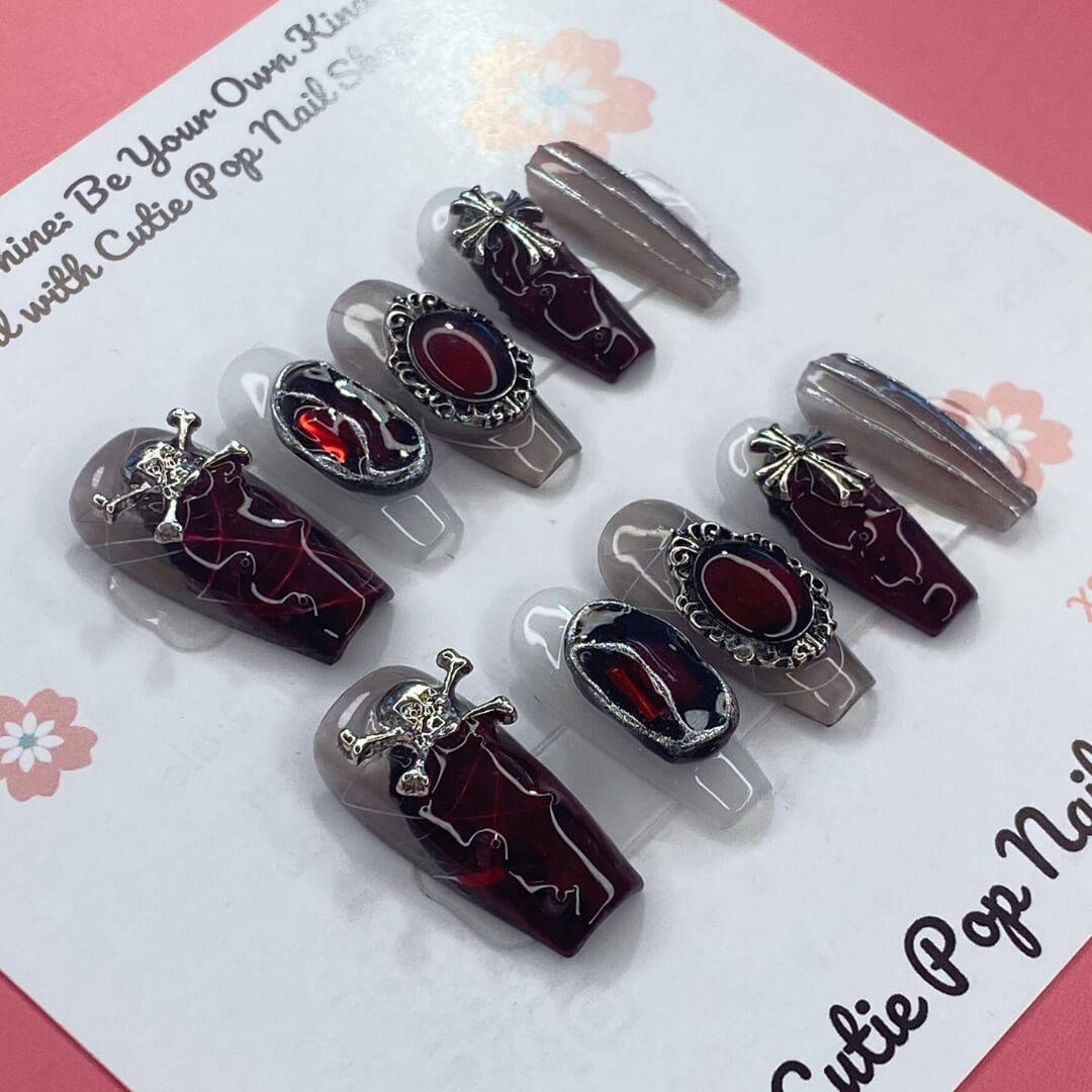 Rebel Chic press‑on nails by CutiePop Nail Shop featuring a medium‑length coffin-shaped set combining glossy jet-black, deep blood-red, and transparent nails accented with edgy skull motifs, mirrored stone embellishments, and stylized cross details—hand-painted and hand-sculpted for a bold, gothic-rock aesthetic.