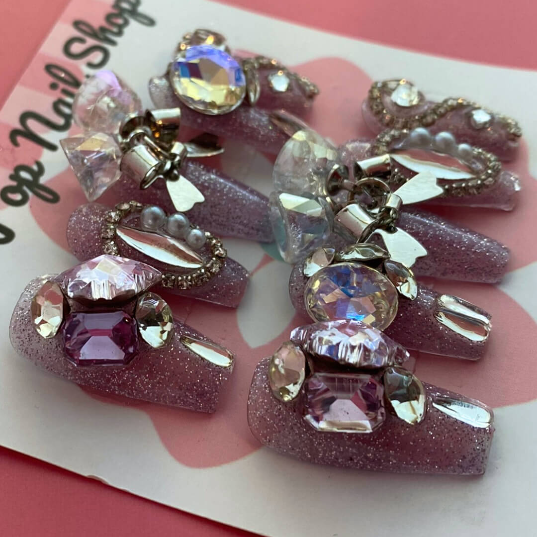 Saturn Bells press‑on nails by CutiePop Nail Shop featuring medium-length coffin‑shaped nails in a deep mauve or dusty rose tone, decorated with oversized iridescent gem clusters, metallic star and planet charms, and delicate glitter accents for a celestial, galaxy‑inspired aesthetic. Includes a set of ten reusable nails, adhesive tabs, mini nail file, and cuticle stick—perfect for dreamy cosmic glam.