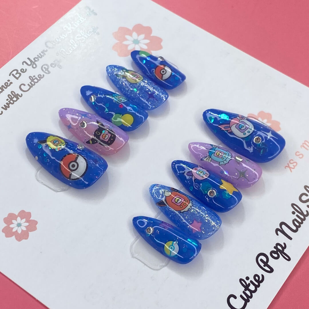 Digipet Evolution press on nails by CutiePop Nail Shop featuring medium almond shaped nails with a glossy royal blue and purple gradient, hand painted with nostalgic anime-style Digipet character silhouettes and sparkly star accents. Includes an easy press on reusable set: adhesive tabs, mini nail file, and cuticle stick.
