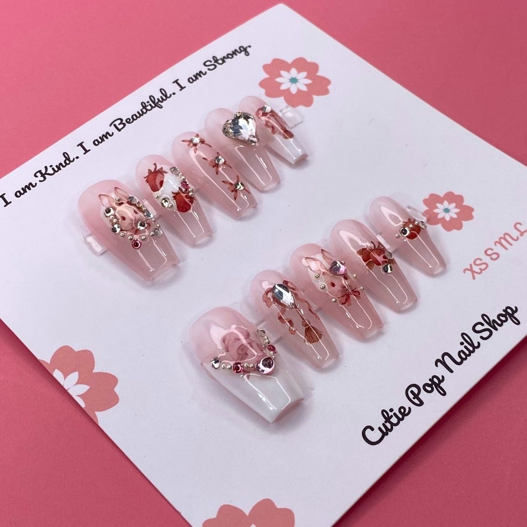 Strawberry Bunny press-on nails by CutiePop Nail Shop featuring medium-length coffin-shaped nails in soft pink tones with adorable strawberry and bunny designs. Accented with tiny bows, pearls, and glossy details for a sweet, kawaii-inspired aesthetic.