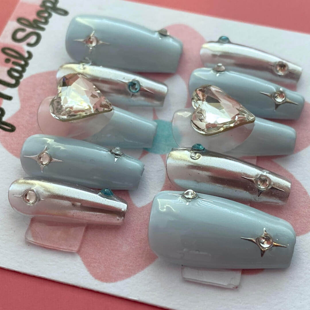 Y2K Blue Heart press‑on nails by CutiePop Nail Shop featuring medium-length coffin-shaped nails in a frosty sky-blue or pastel blue base adorned with bold glossy white or neon blue heart motifs, metallic chrome or holographic shimmer accents, and sparkling rhinestones for a retro Y2K pop-punk aesthetic. Comes with a full kit including 10 reusable handmade nails, jelly adhesive tabs, a mini nail file, and a cuticle stick for easy DIY application.