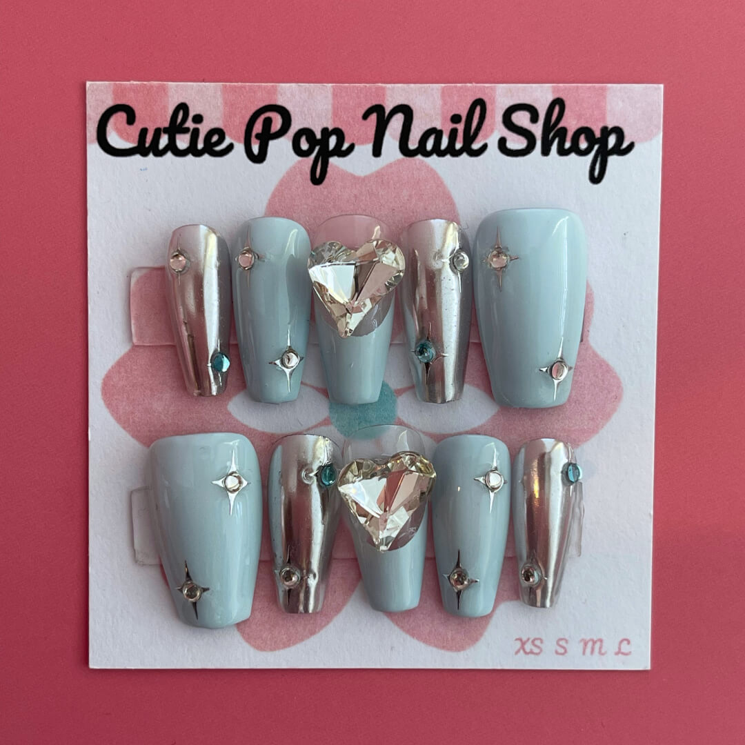 Y2K Blue Heart press‑on nails by CutiePop Nail Shop featuring medium-length coffin-shaped nails in a frosty sky-blue or pastel blue base adorned with bold glossy white or neon blue heart motifs, metallic chrome or holographic shimmer accents, and sparkling rhinestones for a retro Y2K pop-punk aesthetic. Comes with a full kit including 10 reusable handmade nails, jelly adhesive tabs, a mini nail file, and a cuticle stick for easy DIY application.