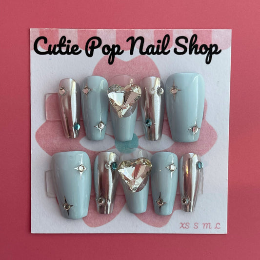 Y2K Blue Heart press‑on nails by CutiePop Nail Shop featuring medium-length coffin-shaped nails in a frosty sky-blue or pastel blue base adorned with bold glossy white or neon blue heart motifs, metallic chrome or holographic shimmer accents, and sparkling rhinestones for a retro Y2K pop-punk aesthetic. Comes with a full kit including 10 reusable handmade nails, jelly adhesive tabs, a mini nail file, and a cuticle stick for easy DIY application.