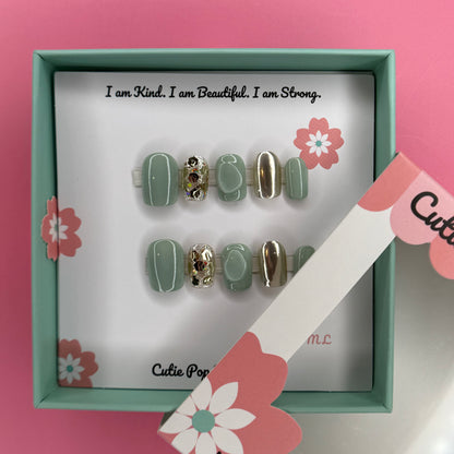Green and Gold Sparkle press-on nails in a rich green base with sparkling gold accents and 3D bump embellishments, finished with a glossy shine – displayed on a Cutie Pop Nail Shop card.
