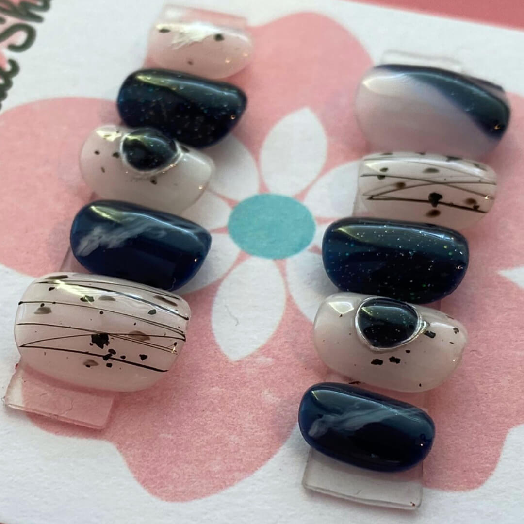Abstract Gem press‑on nails by CutiePop Nail Shop featuring short-length squoval (square-oval) nails in a bold contrasting palette of glossy black and nude tones, accented by irregular abstract brush strokes and oversized metallic gemstone clusters. Includes reusable adhesive tabs, mini nail file, and a cuticle stick for full DIY application—delivering an edgy, artistic manicure with luxe shine.