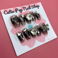 Black Gem Silver Glitter press‑on nails by CutiePop Nail Shop featuring medium-length coffin-shaped nails painted in a deep glossy black base, embellished with a sparkling silver glitter gradient and large gem clusters. The design includes multicolored rhinestones and metallic accents for a dramatic glam‑rock aesthetic. Arrives as a reusable kit—10 handmade press‑on nails, a sheet of jelly adhesive tabs, a mini nail file, and a cuticle stick.

