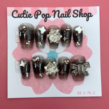 Black Gem Silver Glitter press‑on nails by CutiePop Nail Shop featuring medium-length coffin-shaped nails painted in a deep glossy black base, embellished with a sparkling silver glitter gradient and large gem clusters. The design includes multicolored rhinestones and metallic accents for a dramatic glam‑rock aesthetic. Arrives as a reusable kit—10 handmade press‑on nails, a sheet of jelly adhesive tabs, a mini nail file, and a cuticle stick.

