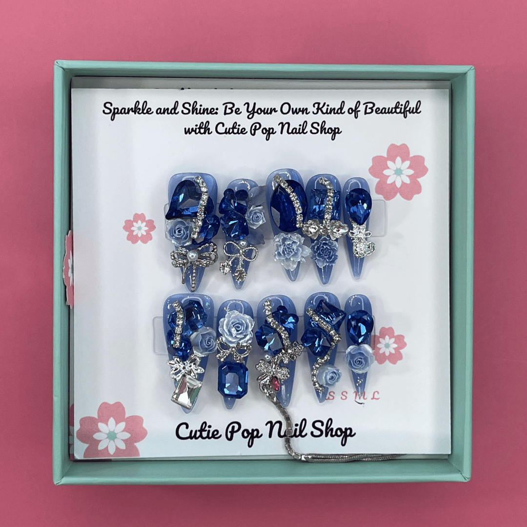 Bougie Bling 7 press on nails by CutiePop Nail Shop featuring long stiletto shaped nails with a vibrant royal blue glitter base adorned with oversized 3D gem clusters in matching blue and clear tones, complemented by sparkling silver rhinestones and metallic accents for a dazzling, high glam luxury statement manicure.

