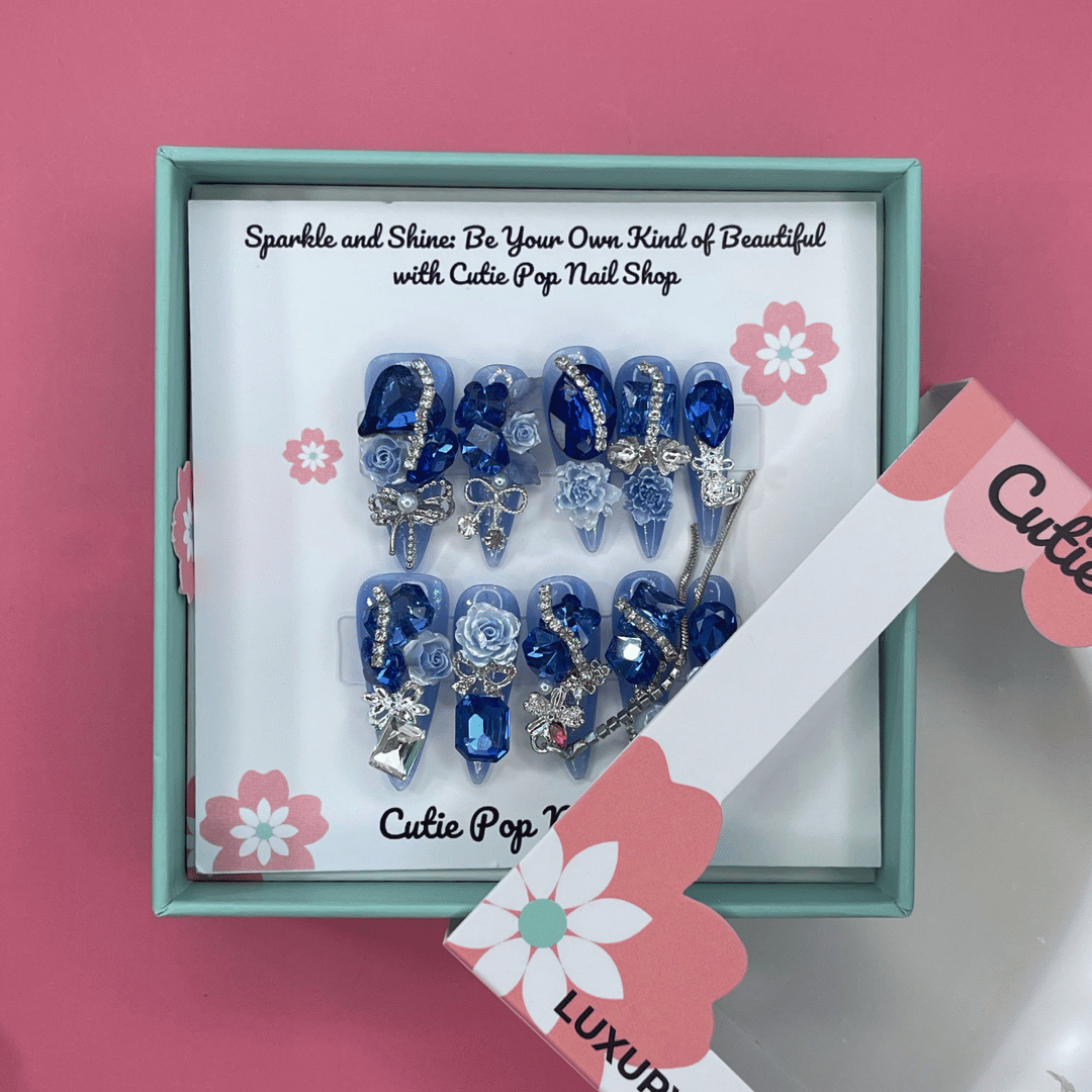 Bougie Bling 7 press on nails by CutiePop Nail Shop featuring long stiletto shaped nails with a vibrant royal blue glitter base adorned with oversized 3D gem clusters in matching blue and clear tones, complemented by sparkling silver rhinestones and metallic accents for a dazzling, high glam luxury statement manicure.

