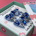 Bougie Bling 7 press on nails by CutiePop Nail Shop featuring long stiletto shaped nails with a vibrant royal blue glitter base adorned with oversized 3D gem clusters in matching blue and clear tones, complemented by sparkling silver rhinestones and metallic accents for a dazzling, high glam luxury statement manicure.

