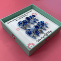 Bougie Bling 7 press on nails by CutiePop Nail Shop featuring long stiletto shaped nails with a vibrant royal blue glitter base adorned with oversized 3D gem clusters in matching blue and clear tones, complemented by sparkling silver rhinestones and metallic accents for a dazzling, high glam luxury statement manicure.

